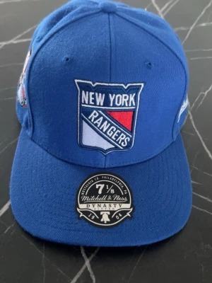 Blå New York Rangers keps - Snygg blå keps från Mitchell & Ness med New York Rangers-logga framtill och ett 75-årsjubileumsmärke på sidan. Klassisk rak skärm och broderade detaljer. Perfekt för dig som gillar hockey och vill ha en sportig stil.