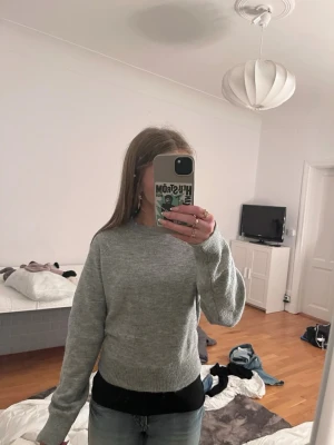 Ljusgrå stickad tröja - En enkel och stilren ljusgrå stickad tröja med rund hals och långa ärmar. Tröjan har ribbade muddar vid ärmslut och nederkant, vilket ger en snygg passform. Perfekt att matcha med jeans eller kjol.