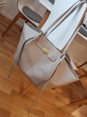 Beige shoppingväska med gulddetalj Gina Tricot - Stilren shoppingväska i beige med långa axelband och snygg guldfärgad spänndetalj framtill. Väskan har en mjuk, slät yta och rymlig form med dekorativa band på sidorna. Perfekt för dig som vill ha plats för allt och ändå se trendig ut.