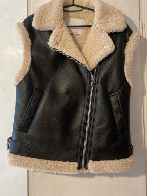 Svart pälsväst med fuskpälsdetaljer - Snygg svart väst i skinnimitation med krage och foder i beige fuskpäls. Västen har dragkedja framtill, två fickor och dekorativa spännen i sidorna. Perfekt för lager på lager och ger en cool look till din outfit.