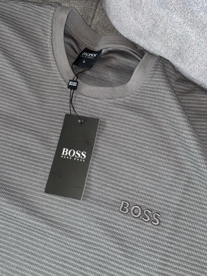 Grå BOSS t-shirt med diskret mönster - Snygg grå t-shirt från BOSS med subtilt randigt mönster och broderad logga på bröstet. T-shirten har rund halsringning och korta ärmar, perfekt för en clean och stilren look. Tillverkad i mjuk bomull för maximal komfort.