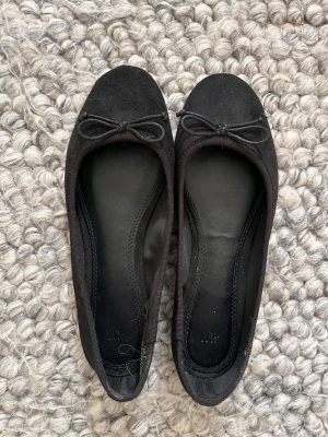 Svarta ballerinaskor med rosett H&M - Stilrena svarta ballerinaskor från H&M med söt rosett framtill. Skorna har rund tå och ovandel i mockaimitation, samt en mjuk innersula för extra komfort. Perfekta för en enkel och klassisk look. Storlek 36