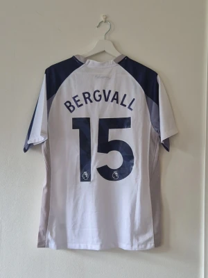 Tottenham matchtröja Bergvall 15 - Säljer en vit Tottenham Hotspur matchtröja från Nike med mörkblå detaljer och grå inslag på sidorna. Tryck med 'Bergvall' och nummer 15 på ryggen, samt AIA-logga på bröstet. Perfekt för fotbollsfans som vill visa sitt stöd på läktaren eller planen.