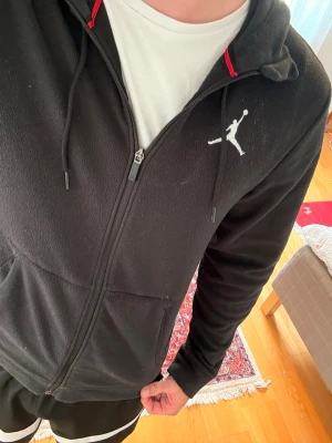 Svart Jordan zip hoodie med huva - Svart hoodie med dragkedja från Jordan, prydd med den klassiska Jumpman-loggan i vitt på bröstet. Hoodien har huva med dragsko och två fickor framtill. Perfekt för en sportig och avslappnad stil. Passar mig som är 191 cm lång men även kortare.