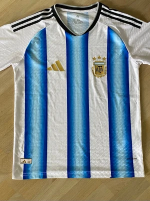 Argentina matchtröja  - Säljer en  Argentina matchtröja från Adidas med klassiska blå och vita vertikala ränder. Tröjan har svarta detaljer vid krage och axlar, guldigt Adidas-märke och AFA-emblem med tre stjärnor. Tillverkad i lätt funktionsmaterial, perfekt för fotboll.