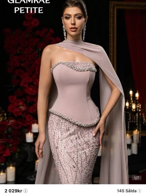 Ljusrosa glittrig långklänning - Elegant ljusrosa långklänning från Glamrae med glittriga detaljer och pärlutsmyckning. Klänningen har en unik asymmetrisk axel med cape, figurnära passform och vackert fall. Perfekt för bal eller festliga tillfällen. Köpt för 2029kr och använd bara en gång. ☺️