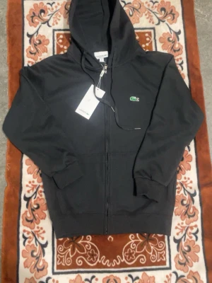 Svart hoodie från Lacoste med dragkedja - Svart hoodie från Lacoste med dragkedja framtill och klassisk grön krokodil-logga på bröstet. Huva med snören och ribbade muddar vid ärmslut och nederkant. Tillverkad i mjukt bomullsmaterial för en skön känsla. Perfekt för en chill och stilren look.
