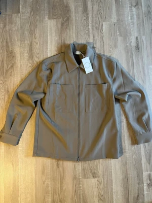 Olivgrön Approach overshirt med dragkedja - Stilren olivgrön overshirt från Approach med två stora bröstfickor och dragkedja framtill. Overshirten har klassisk krage, raka ärmar med knapp vid manschetten och är tillverkad i ett slätt tyg. Perfekt för lager-på-lager och enkel att matcha.