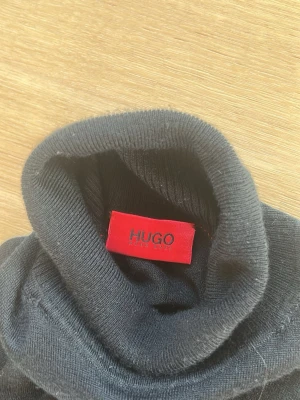 Svart polotröja från HUGO - Svart stickad polotröja från HUGO med klassisk passform och ribbstickad krage. Tröjan har långa ärmar och är tillverkad i ett mjukt, tunt material som passar perfekt för lager-på-lager. Enkel och stilren design som funkar till många outfits.