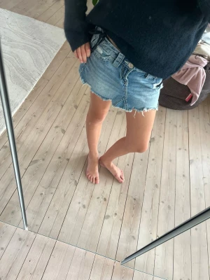 Blå jeanskjol från gina - Säljer en kort blå jeanskjol med rå, fransig kant och slitna detaljer. Kjolen har klassisk femficksmodell och en snygg broderad detalj på bakfickan. Perfekt för dig som gillar en avslappnad och trendig stil. 