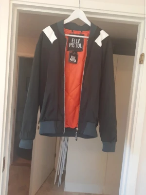 Svart bomberjacka från Elly Pistol - Ny med lappar kvar. Svart bomberjacka från Elly Pistol med orange foder, vita detaljer på axlarna och breda muddar i grått. Jackan har dragkedja framtill med en stor Elly Pistol-zipper och klassisk bomberjacke-look. Perfekt för dig som vill sticka ut med en cool streetstyle.