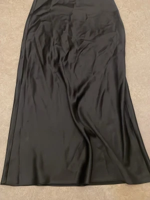 Svart långkjol i satin från Zara - Säljer en svart långkjol från Zara i glansigt satinliknande material. Kjolen har en enkel och stilren design med lätt utsvängd form. Perfekt att styla med en crop top eller stickad tröja för en trendig look.