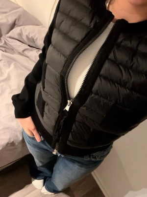Svart dunväst från Moncler - Snygg svart dunväst från Moncler med quiltad front och tvåvägsdragkedja. Västen har stickade svarta ärmar och en Moncler-logga på ena ärmen. Perfekt för lager-på-lager och ger en sportig look. Materialet är en mix av dun och stickat tyg.