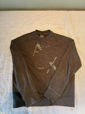 Armani Exchange tröja - Säljer en svart sweatshirt från Armani Exchange med stort tryck på bröstet i metallic och diskret logga. Tröjan har rund hals, långa ärmar och ribbade muddar vid ärmslut och nederkant. Perfekt för en avslappnad och stilren look.