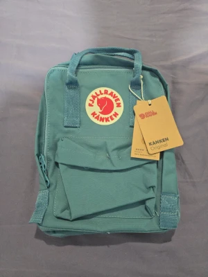 New Blå Fjällräven Kånken ryggsäck - Säljer en klassisk blå Fjällräven Kånken ryggsäck med det ikoniska runda röda logotypmärket framtill. Ryggsäcken har ett stort huvudfack, en mindre ficka framtill och två sidofickor. Tillverkad i slitstarkt material och har både handtag och axelband.