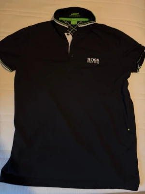 Svart piké från Hugo Boss - Snygg svart pikétröja från Hugo Boss med vit logga på bröstet och coola detaljer i kragen med gröna och vita inslag. Tröjan har knappar framtill och klassisk krage. Perfekt för en stilren och sportig look.