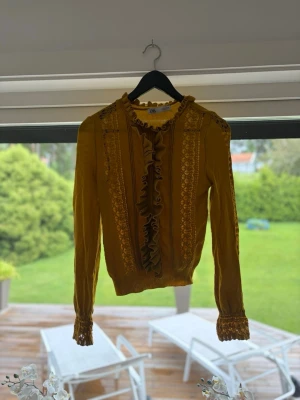 Gul blus med volanger och spets - Trendig gul blus med långa ärmar, volangdetaljer framtill och spets längs ärmar och axlar. Hög hals med liten volangkant och ribbade muddar vid ärmslut. Perfekt för dig som vill sticka ut med en färgglad och detaljerad look.