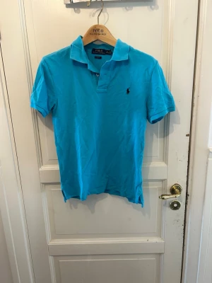 Blå pikétröja från Polo Ralph Lauren - Snygg blå pikétröja från Polo Ralph Lauren i slim fit-modell. Klassisk krage, korta ärmar och den ikoniska loggan broderad på bröstet. Perfekt till jeans eller shorts för en clean och fräsch look.
