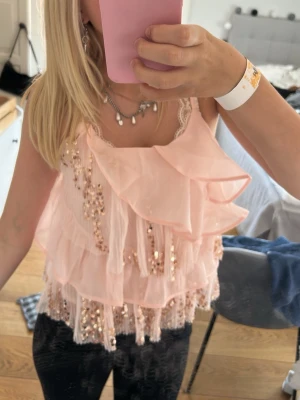 Ljusrosa volangtopp med paljetter - Supersöt ljusrosa topp med tunna axelband, flera lager volanger och glittriga paljettdetaljer. Toppen är ärmlös och har en luftig, flowy passform som känns riktigt trendig. Perfekt för dig som vill sticka ut lite extra med en feminin och lekfull stil.