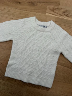 Vit kabelstickad tröja - Kabelstickad vit tröja med rund hals och ribbade muddar. Tröjan har ett klassiskt flätmönster framtill och är gjord i ett mjukt material som känns skönt mot huden. Perfekt för kyliga dagar och enkel att matcha med olika outfits.