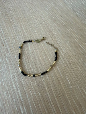 Svart och guldigt pärlarmband - Snyggt armband med små svarta och guldfärgade pärlor i ett enkelt men trendigt mönster. Armbandet har en tunn kedja och ett metallspänne i guldton. Perfekt accessoar för att lyfta din outfit med en stilren touch.