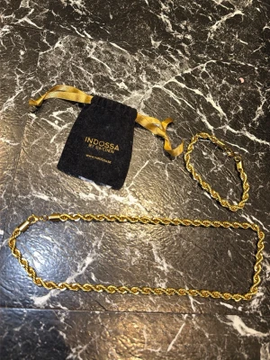 Guldarmband från Indossa - Snyggt guldarmband från Indossa med tvinnad kedja i metall. Armbandet har en elegant och stilren look och kommer med en svart tygpåse med guldfärgade band och logga. Perfekt accessoar för att lyfta din outfit.