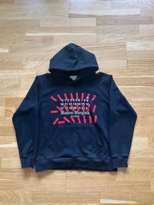 Maison Margiela hoodie - Svart hoodie från Maison Margiela med ikoniskt tryck framtill i vitt och röda detaljer. Storlek S. Klassisk känguruficka och huva.