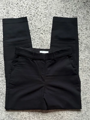 Svarta raka kostymbyxor - Stilrena svarta kostymbyxor med raka ben och klassisk passform. Byxorna har hällor för skärp, sidofickor och en bakficka. Perfekta för en clean och snygg look. Materialet ser ut att vara en mjuk och lite stretchig bomullsblandning.