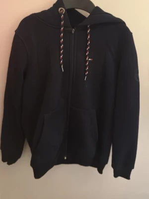 Svart hoodie från Tommy Hilfiger - Snygg svart hoodie från Tommy Hilfiger med dragkedja framtill, två fickor och justerbar huva med mönstrade snören i rött, vitt och blått. Liten logga på bröstet och diskret patch på ärmen. Perfekt för en chill och stilren look.