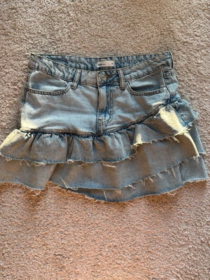 Kort jeanskjol med volanger - Snygg ljusblå jeanskjol med två lager volanger och råa kanter. Kjolen har klassiska fickor fram och bak samt bälteshällor. Perfekt för dig som vill ha en cool och trendig denimlook.