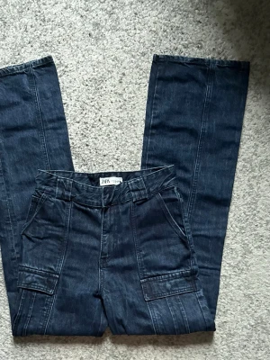 Mörkblå raka jeans från Zara - Säljer ett par mörkblå jeans från Zara med raka ben och coola sömdetaljer framtill. Jeansen har stora fickor både fram och bak samt bälteshällor. Materialet är klassisk denim i bomull. Perfekta för en avslappnad och trendig look.