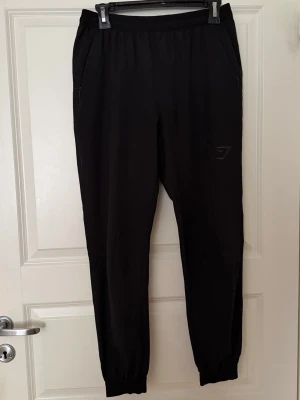 Svarta träningsbyxor från Gymshark - Säljer ett par svarta träningsbyxor från Gymshark med bra passform och resår i midjan samt vid bensluten. Byxorna har dragkedjeförsedda fickor på sidorna och är tillverkade i ett mjukt, stretchigt material som passar perfekt för gym eller löpning.