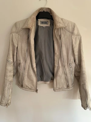 Offwhite/benvit skinnjacka från Mission - Snygg beige skinnjacka från Mission med flera dragkedjor framtill och på ärmarna. Jackan har en klassisk krage och figurnära passform som ger en cool look. Perfekt för dig som vill ha en stilren och trendig jacka till din outfit.