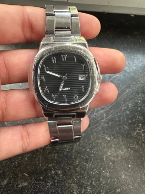Quartz Klocka  - Stilren armbandsklocka i silverfärgat metall med svart urtavla och arabiska siffror. Klockan har datumvisning och ett klassiskt kvartsurverk. Länken är i borstat stål och ger en tidlös look som passar till många stilar.