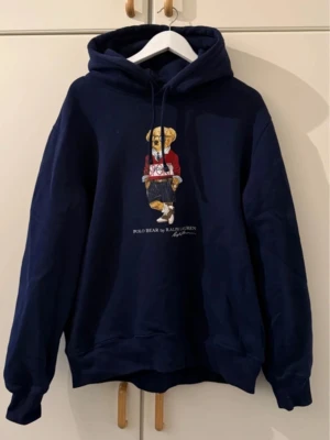 Mörkblå hoodie Polo Bear Ralph Lauren - Mörkblå hoodie från Ralph Lauren med Polo Bear-tryck på bröstet. Klassisk modell med huva och dragsko, samt ribbade muddar vid ärmslut och nederkant. Använd fåtal gånger. Inköp på NK i Stockholm.