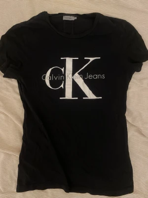 Svart Calvin Klein Jeans t-shirt - Snygg svart t-shirt från Calvin Klein Jeans med stort vitt CK-tryck på bröstet. Klassisk rund halsringning och korta ärmar. Tillverkad i mjuk bomull för en bekväm känsla. Perfekt för en stilren outfit 