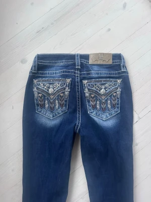 Miss me jeans - Säljer ett par blå jeans från Miss Me med snygga broderade bakfickor och dekorativa detaljer. Jeansen har kontrastsömmar, normal passform och coola nitar på fickorna. Perfekta för dig som vill sticka ut lite extra! Storlek w25. Jeansen är bootcut och midwaist🥰