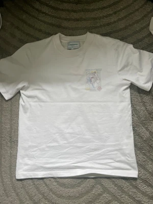 Vit Casablanca t-shirt med grafiskt tryck - Snygg vit t-shirt från Casablanca med ett unikt grafiskt tryck i flera färger på bröstet och ryggen. T-shirten har rund hals och korta ärmar, tillverkad i mjuk bomull för skön känsla. Perfekt för dig som gillar stilrena plagg med en twist.