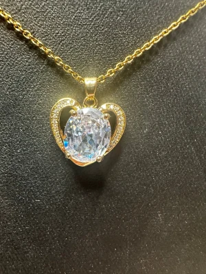 Guldhalsband med stor glittrig sten - Snyggt halsband i guldfärg med en stor, oval glittrig sten i mitten. Hänget har en hjärtformad design med små gnistrande stenar längs kanterna för extra bling. Perfekt för dig som vill sticka ut och addera lite lyx till din outfit.