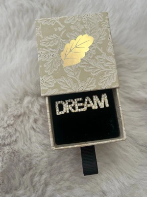 Statementring DREAM i silverfärg - Cool statementring i silverfärg med texten DREAM, täckt av gnistrande stenar som ger en riktigt glittrig look. Ringen har en rak och bred form som verkligen syns och passar perfekt för dig som gillar att sticka ut med dina accessoarer.