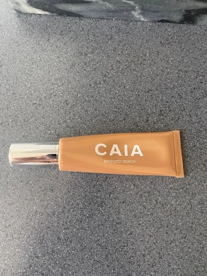 CAIA Bronzed Beach Liquid Highlighter - Flytande highlighter från CAIA i nyansen Bronzed Beach. Kommer i en mjuk tub med silvrigt lock och ger en varm, bronzig glow till huden. Perfekt för att framhäva ansiktets höga punkter med en solkysst finish. Endast testad