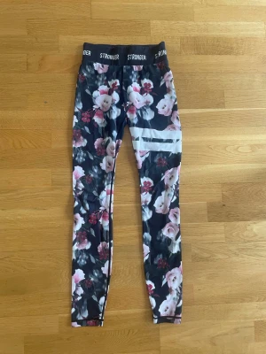 Blommiga leggings från Stronger XS - Snygga leggings från Stronger i storlek XS med svart botten och blommigt mönster i rosa, vitt och rött. Hög midja med elastisk linning där det står 'STRONGER'. Tillverkade i 85% polyester och 15% elastan, perfekta för träning eller chill. Pris kan diskuteras.