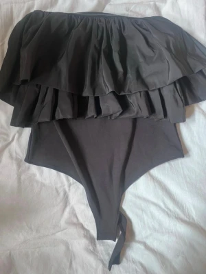 Svart off shoulder body med volanger - Snygg svart body med off shoulder-design och dubbla volanglager upptill. Tillverkad i mjukt, stretchigt material som sitter skönt mot kroppen. Perfekt för dig som vill ha en trendig och stilren look. Knäppning i grenen för enkel på- och avtagning.