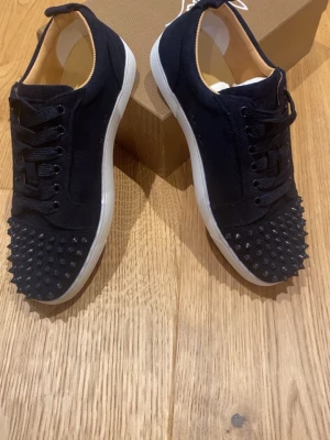 Svarta sneakers med nitar - Snygga svarta sneakers med vita sulor och coola svarta nitar över tån. Skorna har snörning och är tillverkade i mockaliknande material. Röd detalj under sulan ger extra edge till looken.