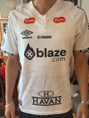 Vit Umbro fotbollströja Neymar  - Säljer en vit fotbollströja från Umbro med flera tryck, NEYMAR I Santos Tröjan har korta ärmar med svartvitrandiga detaljer vid ärmslut och tryck på både bröst och ärmar. Perfekt för dig som gillar sportig stil. Logan har gått av