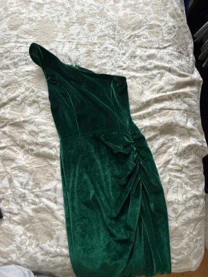 Grön sammetsklänning oneshoulder - Stilren långklänning i mörkgrön sammet med oneshoulder-design och draperad detalj på sidan. Klänningen har en elegant siluett och passar perfekt för speciella tillfällen.