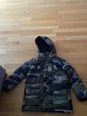 Kamo mönstrad dunjacka med huva - Säljer en camo-mönstrad dunjacka med huva och dragkedja framtill. Jackan har två stora fickor med lock och tryckknappar samt en bröstficka med dragkedja. Muddar vid ärmslut för extra värme. Perfekt för kalla vinterdagar.