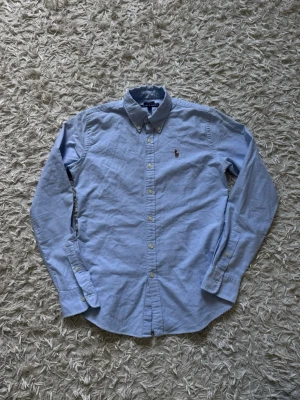 Ljusblå skjorta | Ralph Lauren - Klassisk ljusblå skjorta från Ralph Lauren med broderad logga på bröstet. Skjortan har knappar framtill, button-down krage och långa ärmar. Tillverkad i mjuk bomull för en bekväm känsla och stilren look. I princip helt oanvänd! 