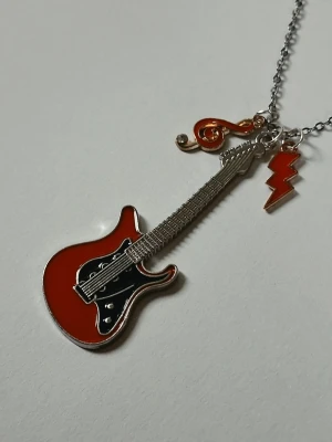 Halsband med elgitarr och berlocker - Unikt halsband med en röd och svart elgitarr i metall som hänge, kompletterat med en röd blixt och en röd notberlock. Kedjan är silverfärgad och detaljerna ger ett coolt och rockigt intryck. Materialet rostfritt stål 