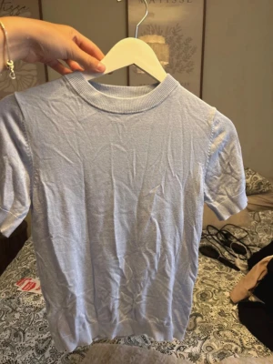 Ljusblå stickad kortärmad topp - En enkel och stilren ljusblå stickad topp med korta ärmar och rund halsringning. Toppen har ribbade kanter vid hals, ärmslut och nederkant. Perfekt att matcha med jeans eller kjol för en fräsch look.